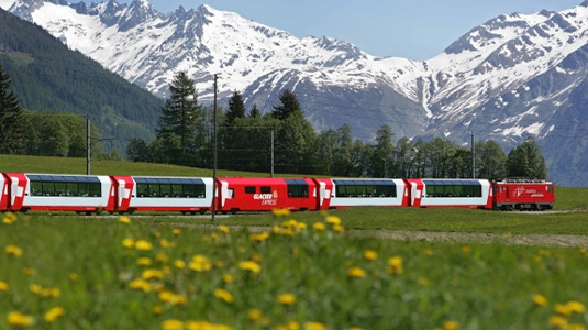 Glacier Express Train running with a green field and mountains in the background.