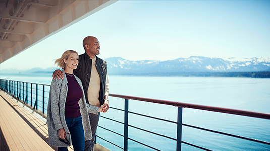 Couple walking hand in hand on a cruise ship deck with water and mountains in the background